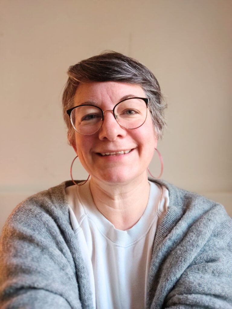 Foto: Mimi lächelt in die Kamera. Sie trägt eine graue Strickjacke über einem hellen Sweatshirt.